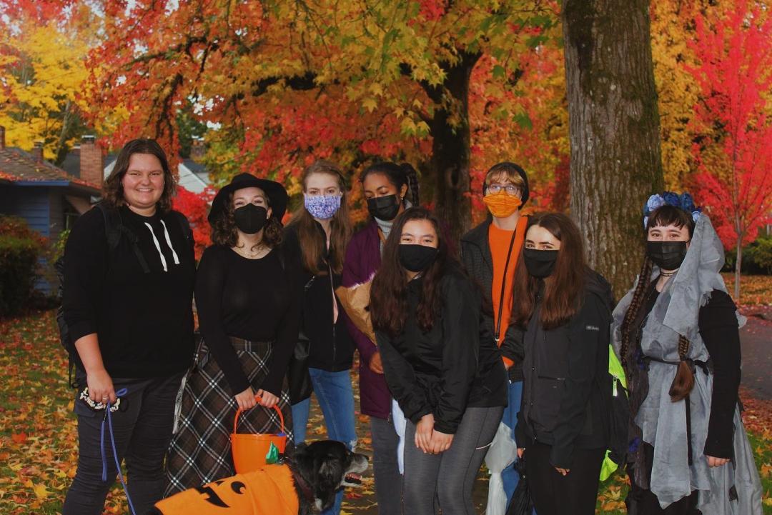 A group of students dressed in dark shirts and jackets, jeans and a few Halloween costumes gather beneath vibrant fall trees. They stand on the sidewalk of a street lined with houses and lawns that are covered in leaves. A black dog with a graying muzzle sits in front of the group wearing a jack-o-lantern costume.