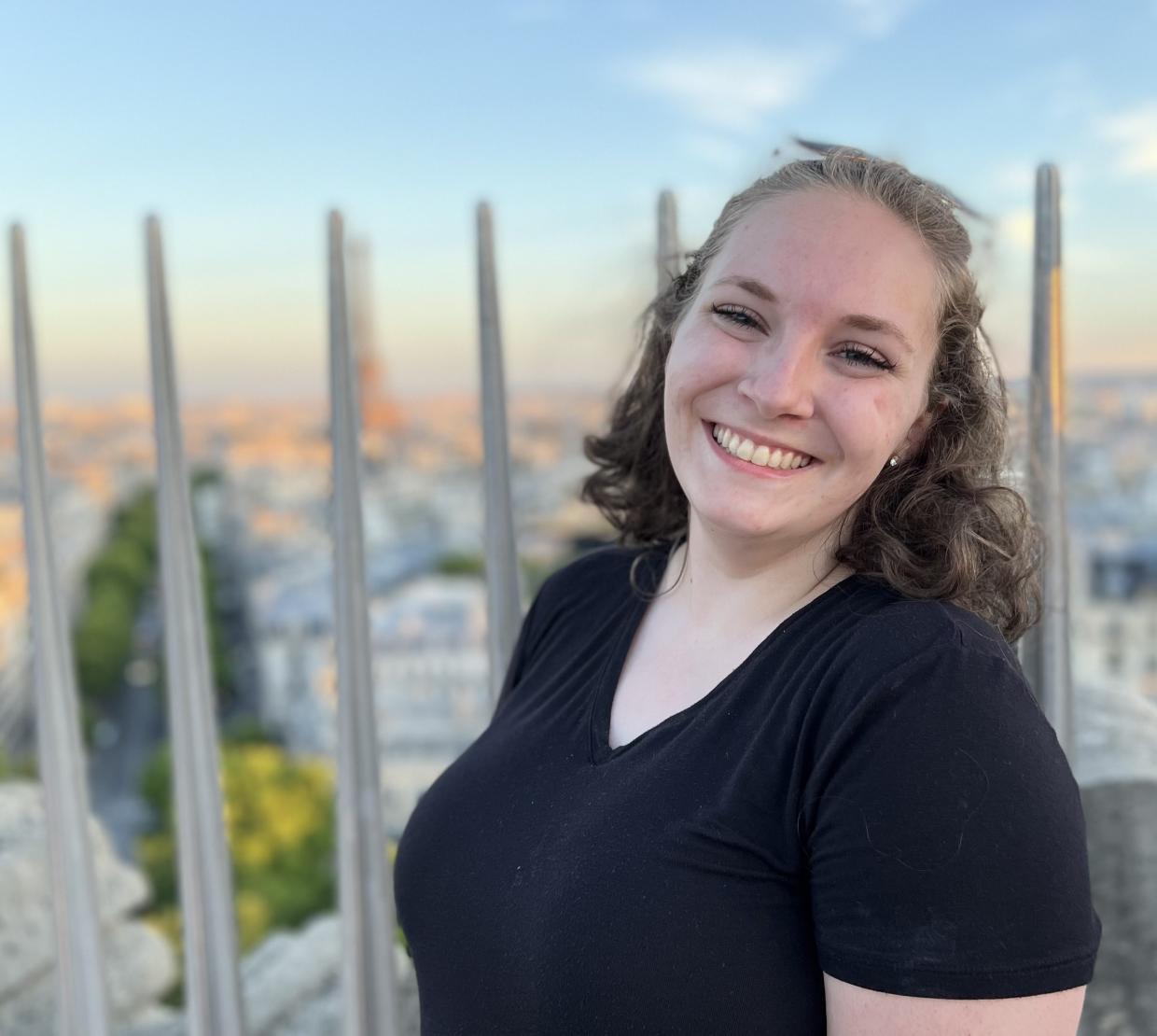 A woman with a dark t-shirt and pulled back hair smiles widely at the camera, the cityscape of Paris blurred in a sunset behind her.