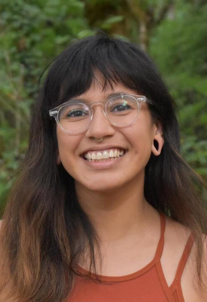 Headshot of Caroline Hernandez outside on an overcast day infront of dark green foliage. She is wearing an orange-red tank top and clear-framed glasses.